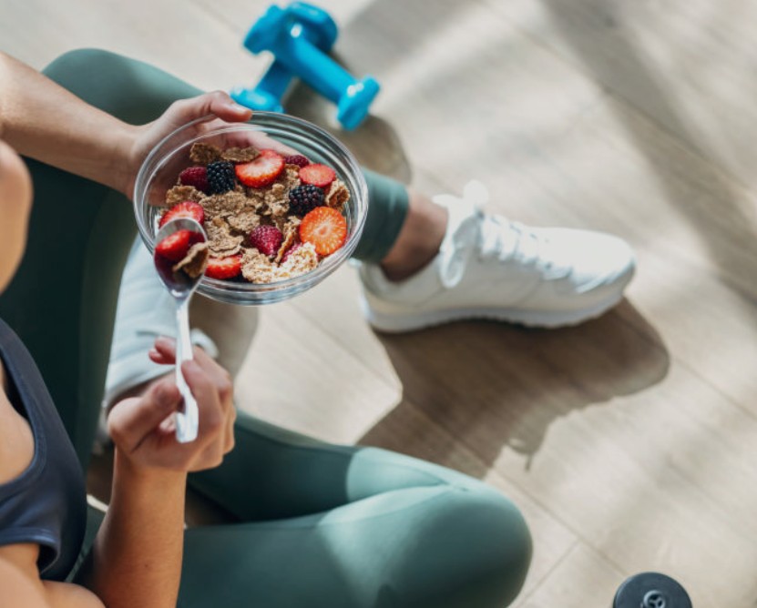 Persona seduta a terra con abbigliamento sportivo che mangia una ciotola di cereali con frutti di bosco dopo l'attività fisica.