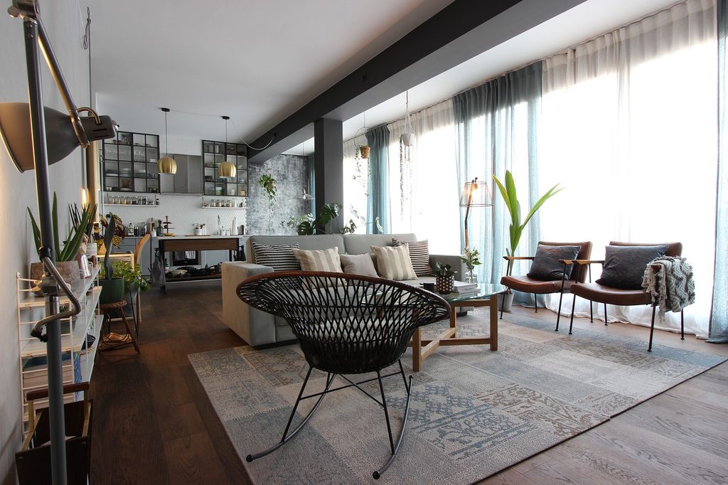 Interno di un open space moderno con zona living e cucina a vista, caratterizzato da pavimenti in legno, ampie vetrate con tende e arredi contemporanei.