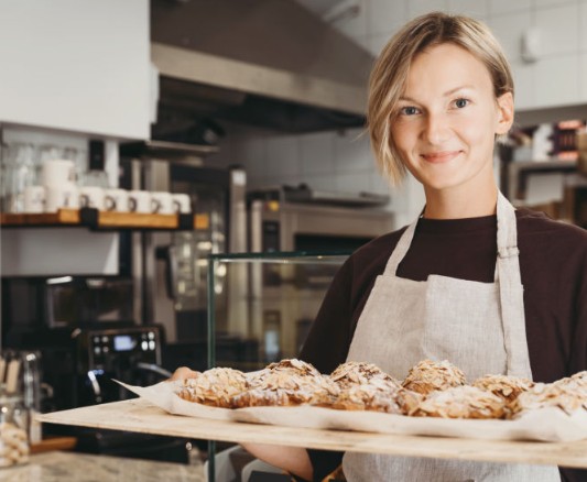 Una donna sorridente indossa un grembiule mentre porge un vassoio di cornetti alle mandorle in una pasticceria.