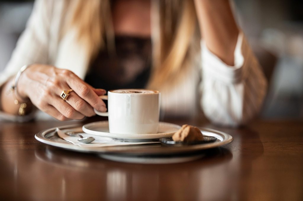 Una persona con una giacca chiara tiene in mano una tazza di caffè su un tavolino di legno in un locale.