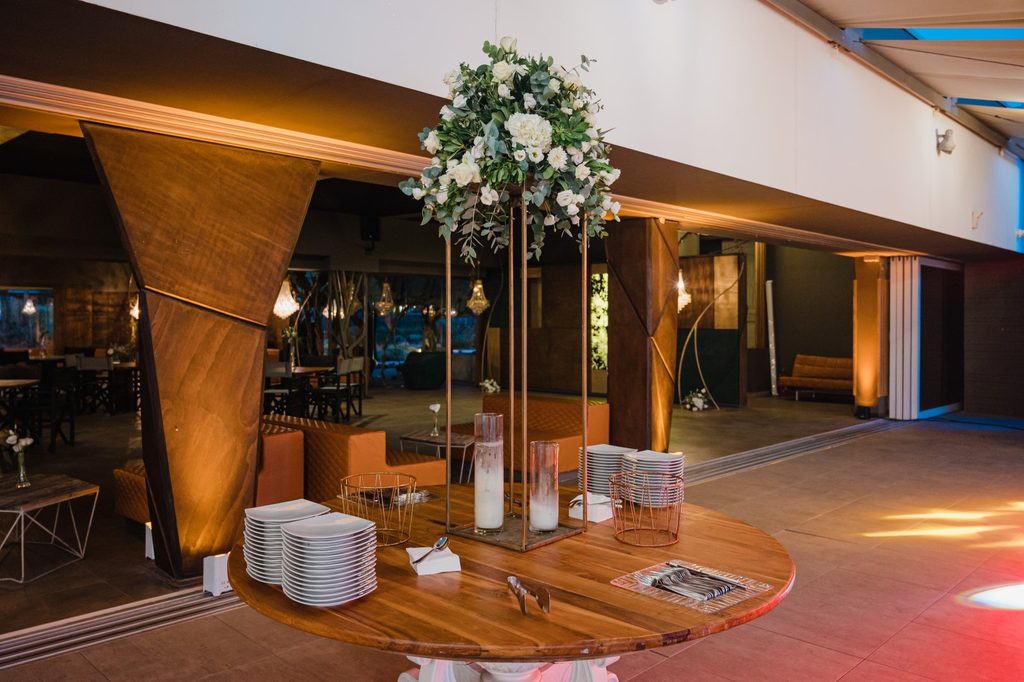 Tavolo rotondo in legno apparecchiato con pile di piatti bianchi e centrotavola floreale in un locale dal design contemporaneo.
