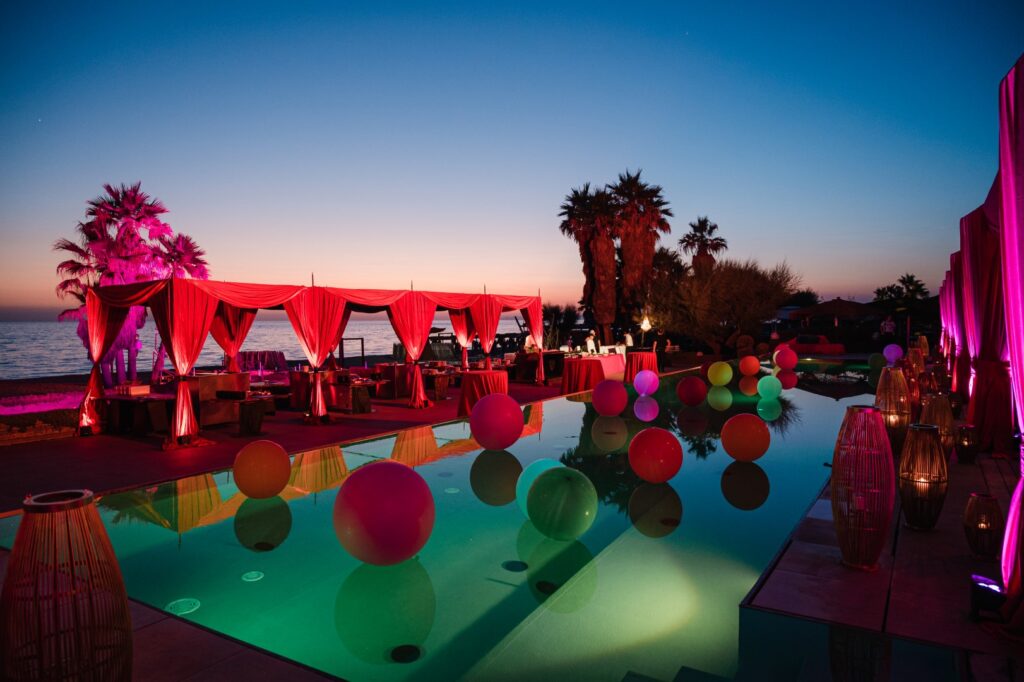 Allestimento scenografico con gazebo rossi e sfere luminose galleggianti in una piscina al tramonto vicino al mare.