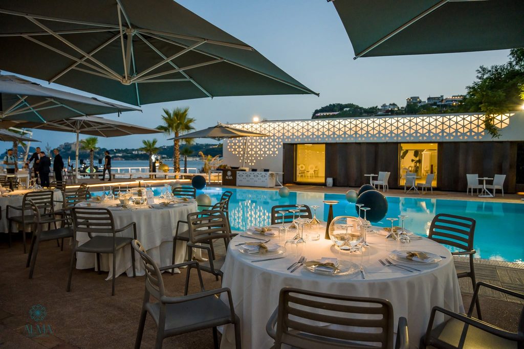 Tavoli apparecchiati per la cena a bordo piscina presso Alma Beach al tramonto.