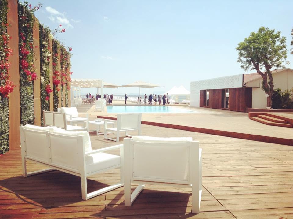 Area lounge esterna con divani bianchi su pedana in legno, piscina e parete vegetale presso Alma Beach a Pozzuoli.