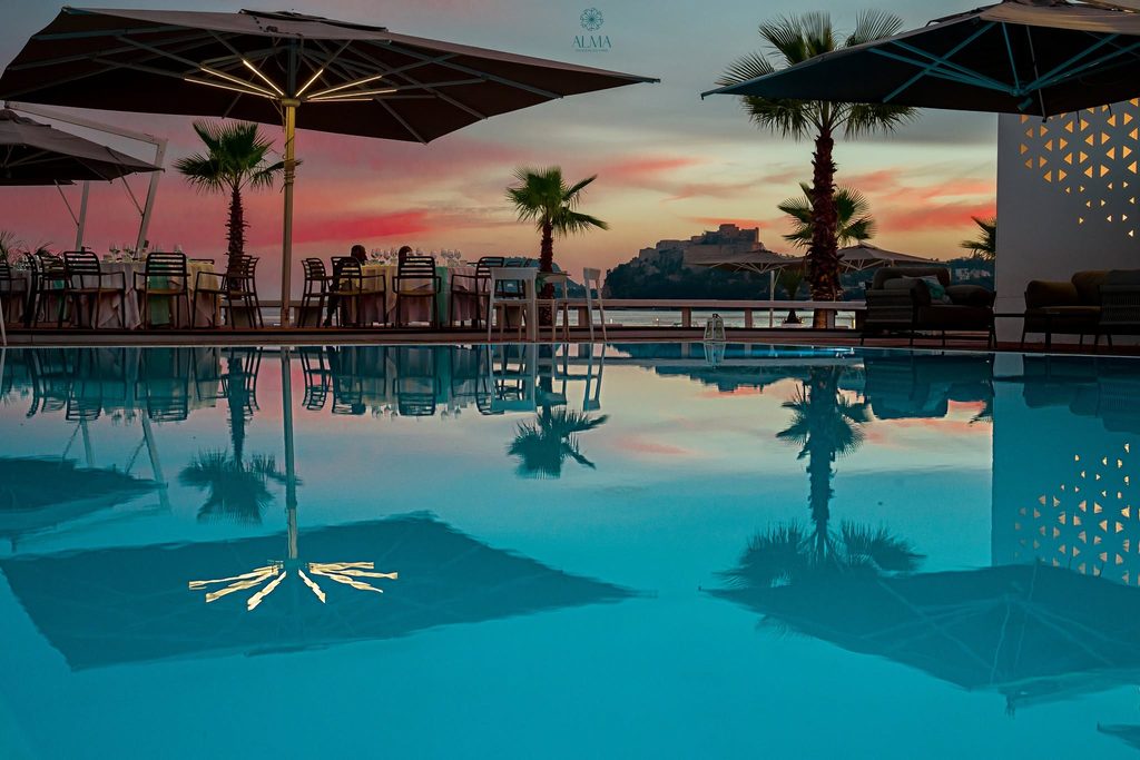 Vista panoramica di una terrazza con piscina al tramonto presso Alma Beach a Pozzuoli, con tavoli apparecchiati e il Castello Aragonese sullo sfondo.