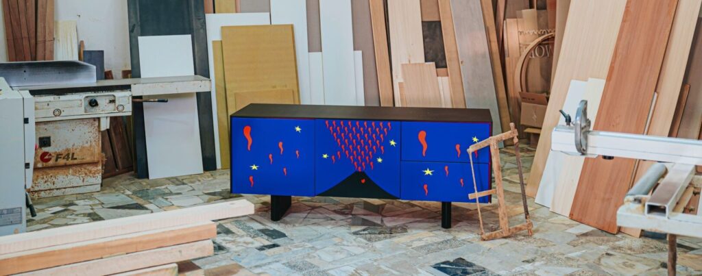 Una madia blu con decorazioni a forma di vulcano in eruzione, stelle gialle e forme rosse, posizionata in un laboratorio di falegnameria.